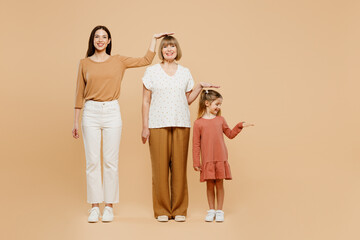 Wall Mural - Full body smiling women wearing casual clothes with child kid girl 6-7 years old. Granny mother daughter show height hold hand above head isolated on plain beige background. Family parent day concept.