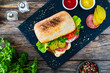 Ciabatta  sandwich with salami, mozzarella, lettuce, tomato, cucumber and dressing on wooden table
