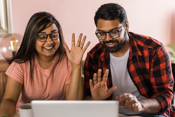 Happy indian family couple cuddle at desk make video call to friends using laptop webcam. Loving young spouses look at computer screen waving hands in good mood greeting parents communicating online