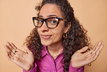 Beautiful Doubtful Curly Brazilian Woman Spreads Palms Has No Idea Or Clue Looks With Indecisive Expression Cannot Make Decision Wears Spectacles And Lila Shirt Poses Against Brown Generative AI