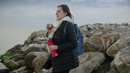 Wall Mural - Young woman relaxing by the sea, holding a thermos of hot tea and admiring the wild and rugged coastline. Ideal for use in tourism, travel, or lifestyle content