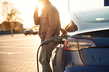 Wall Mural - Talking by phone. Man is standing near his electric car outdoors