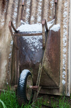 Old Rusty Metal Wheelbarrow With Snow In Front Of A Wall