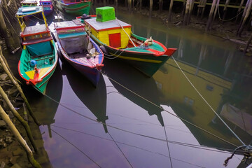 Many small passenger ships is at anchor between wooden buildings in shallow water. Colorful fishing boats in harbor during the day. Small engine boats.