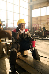 Wall Mural - Hungry worker of industrial factory having homemade sandwich with cup of tea while sitting on wooden box in front of camera in workshop