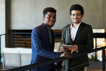 Poster - Two happy young intercultural male brokers in formalwear looking at camera while one of them using tablet to work with online data