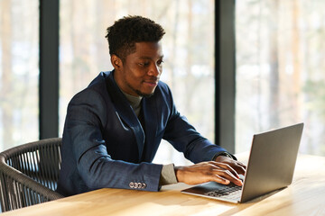 Poster - Young smiling African American entrepreneur typing on laptop keyboard while sitting by workplace in office and working over new project