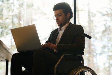 Wall Mural - Young multi-ethnic businessman with disability using laptop while sitting in wheelchair in front of camera and preparing new project