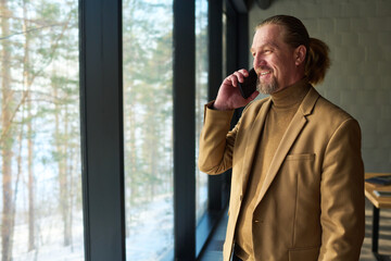 Poster - Smiling chief executive officer in formalwear looking through large office window while standing in front of camera and talking on smartphone