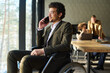 © pressmaster - Young confident businessman in whellchair sitting in front of window and consulting client on smartphone against coworkers having meeting