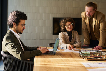 Poster - Side view of young businessman typing on laptop keyboard by workplace against two mature coworkers discussing financial documents