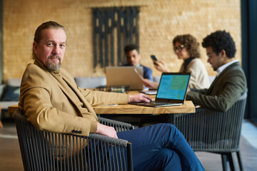 Poster - Confident mature director of business company sitting by workplace in front of laptop and looking at camera against his subordinates