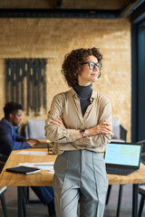Aufkleber - Young pretty business leader in quiet luxury attire looking through window while standing in front of camera by workplace against coworker