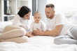 © sofiko14 - Caucasian parents lying on bed with baby and tenderly embracing from both sides. Sweet little girl with funny ponytail smiling lightly and looking ahead. Concept of love and family treasures.