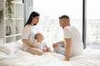 © sofiko14 - Cute little baby girl sitting together with loving parents on soft duvet in bedroom during weekend at home. Cheerful caucasian mom and dad taking care of pretty daughter while relaxing indoors.