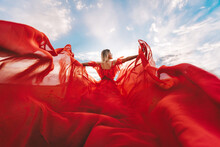 Woman Red Flying Dress. A Blonde In A Red Dress Against The Sky. Rear View Of A Beautiful Blonde Woman In A Red Dress Fluttering In The Wind Against A Blue Sky And Clouds.