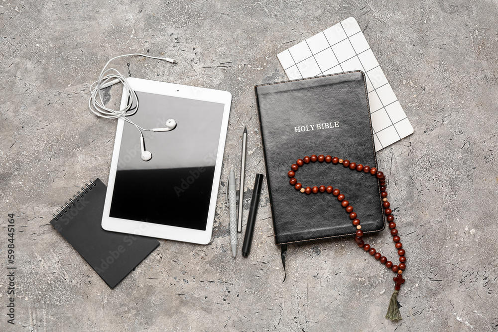 Holy Bible with prayer beads, notebooks and tablet computer on grunge background