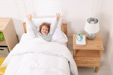 Sticker - Cute little boy stretching in cosy bed near alarm clock on bedside table indoors