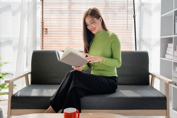 Asian young woman making notes in paper notebook sitting on cozy sofa on background of window. Pretty female writing notes in diary or making to do list relaxing at home.