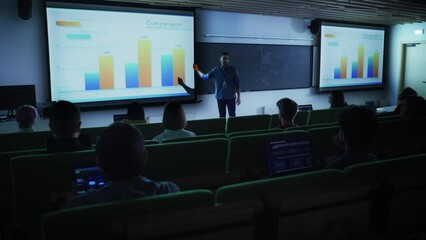 Fototapete - Young Male Teacher Giving a Business Strategy Lecture to Diverse Multiethnic Group of Female and Male Students in Dark College Room. Projecting Slideshow with Comparison Charts and Commercial Data