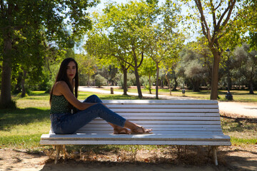 beautiful Spanish woman with black hair is sitting on a white bench in the park. She is alone and pensive. Concept expressions and gestures.