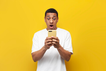 young shocked african american man in white t-shirt is using smartphone and surprised on yellow isolated background