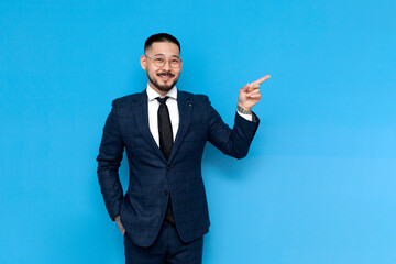 Wall Mural - successful asian businessman in suit pointing with hand at copy space on blue isolated background