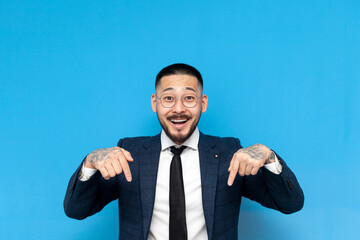 successful asian businessman in suit and glasses points down with his hands on blue isolated background