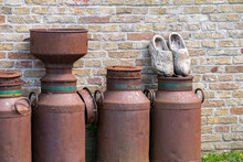 Dutch Traditional Wooden Clogs And Old Rusty Shabby Milk Cans In A Vintage Farmhouse Decoration. Vintage Composition In Rural House Style.