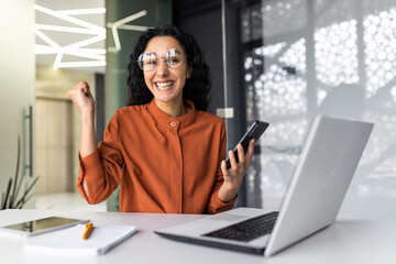 Wall Mural - Portrait of successful Latin American business woman, worker in glasses and curly hair looking at camera and smiling happy celebrating triumph and winning success sitting at table holding phone.