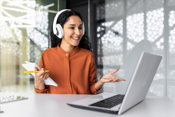 Wall Mural - Young beautiful and successful business woman studying remotely sitting in the office, Hispanic woman writing in notebook using laptop for video call, talking with colleagues and mentor remotely