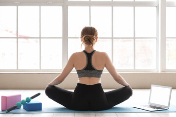 Poster - Sporty young woman with laptop doing yoga in gym, back view