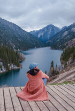Girl Sits On The Pier And Looks At The Kolsai Lake And Mountains. Travel Concept In Kazakhstan. High Quality Vertical Photo.