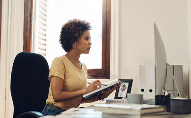 Poster - Working tirelessly to reach success. a young businesswoman working on a computer while going through notes from a book in an office.