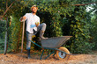 © _KUBE_ - Organic gardening. Handsome caucasian bearded gardener poses with his foot on a cart holding pitchfork. The concept of summer and harvesting