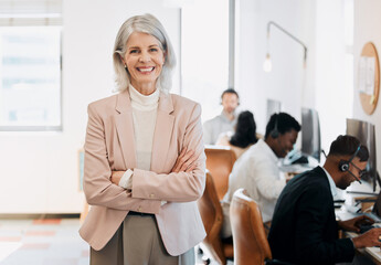 Im in charge. a mature businesswoman standing in the office with her arms folded while her team work behind her.