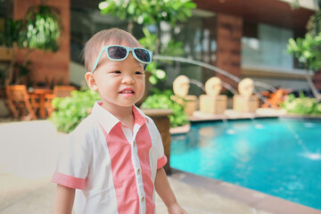 Portrait of cute little Asian 2 years old toddler boy child with sunglasses wearing stylish shirt smiling near the pool at hotel in the summer outdoors, Happy summer vacation for children concept