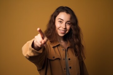 Wall Mural - Portrait of smiling young woman pointing finger at camera over brown background, Generative AI