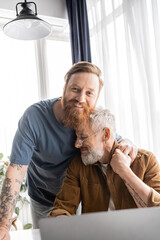 Wall Mural - Carefree homosexual man holding hand of partner near blurred laptop at home. 