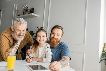 Wall Mural - Smiling gay parents with daughter looking at camera while doing homework at home. 