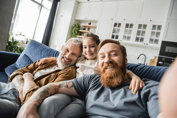 Wall Mural - Smiling preteen kid hugging same sex parents and looking at camera on couch at home. 