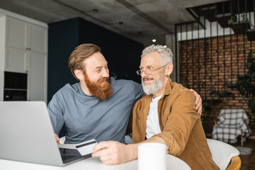 Wall Mural - Positive gay man hugging partner with credit card near laptop and coffee at home. 