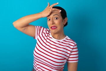 Sticker - Oops, what did I do? beautiful woman wearing striped T-shirt over blue studio background  holding hand on forehead with frightened and regret expression.