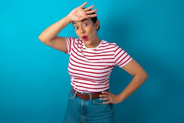 Wall Mural - beautiful woman wearing striped T-shirt over blue studio background wiping forehead with hand making phew gesture, expressing relief feels happy that he prevented huge disaster. It was close enough