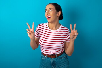 Wall Mural - Isolated shot of cheerful beautiful woman wearing striped T-shirt over blue studio background makes peace or victory sign with both hands, feels cool.