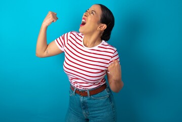 Sticker - Attractive beautiful woman wearing striped T-shirt over blue studio background celebrating a victory punching the air with his fists and a beaming toothy smile