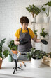 © satura_ - Relaxing home gardening. Smiling middle aged woman in black gloves with potted plant records gardening video blog in modern house - blogging and florist vlog influencer