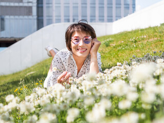 Wall Mural - Woman in colorful sunglasses sniffs clover flowers on lawn in urban park. Nature in town. Relax outdoors after work. Summer vibes.