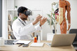© sofiko14 - Side view of multiethnic male in white coat using virtual reality headset while sitting at office desk with digital gadgets near anatomical model. Professor simulating medical situation in clinic.