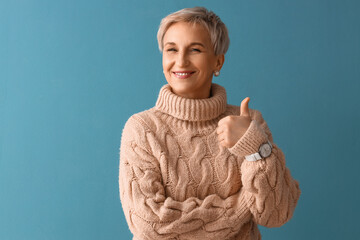 Poster - Beautiful mature woman in beige sweater showing thumb-up on blue background